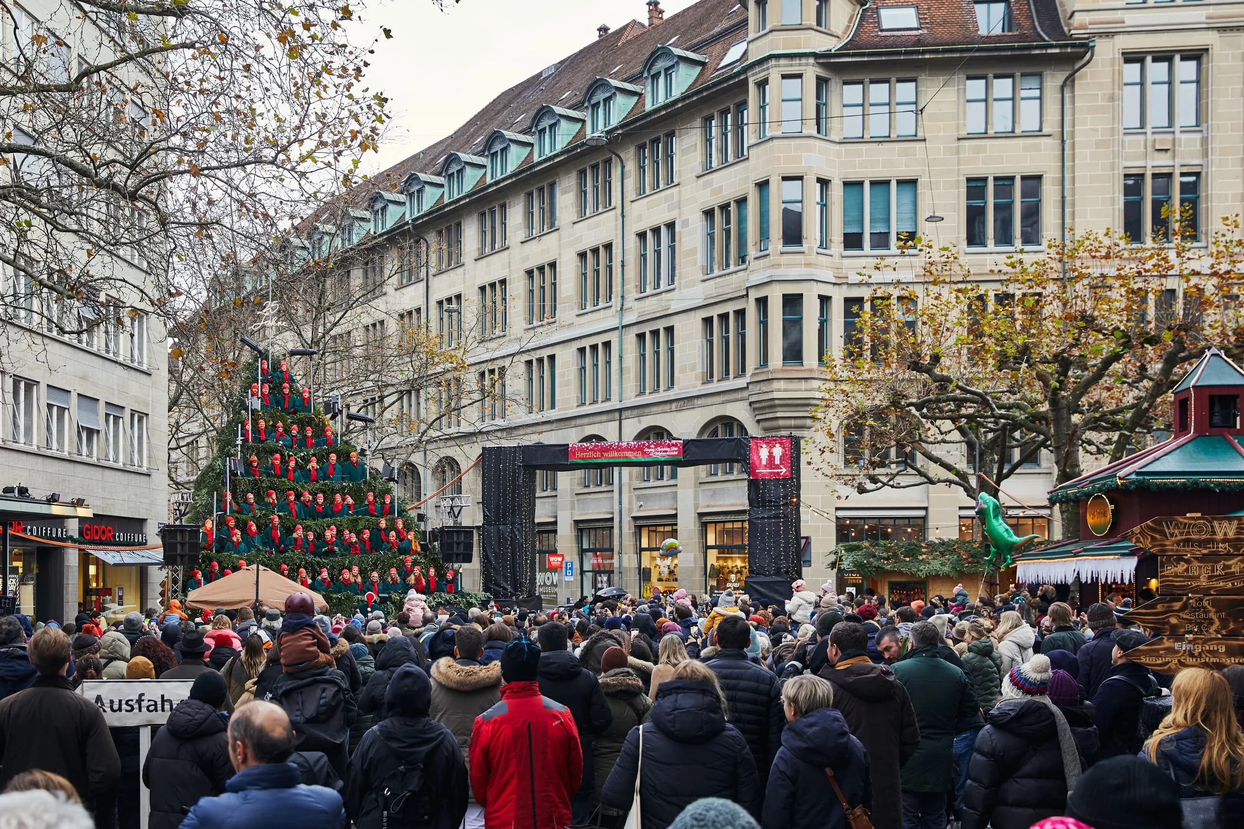 Grosses Publikum in Winterjacken steht tagsüber auf dem Werdmühleplatz vor dem Singing Christmas Tree mit Leuchtschrift «25 Jahre», daneben der Eingangsbogen zum Wiehnachtsmärt