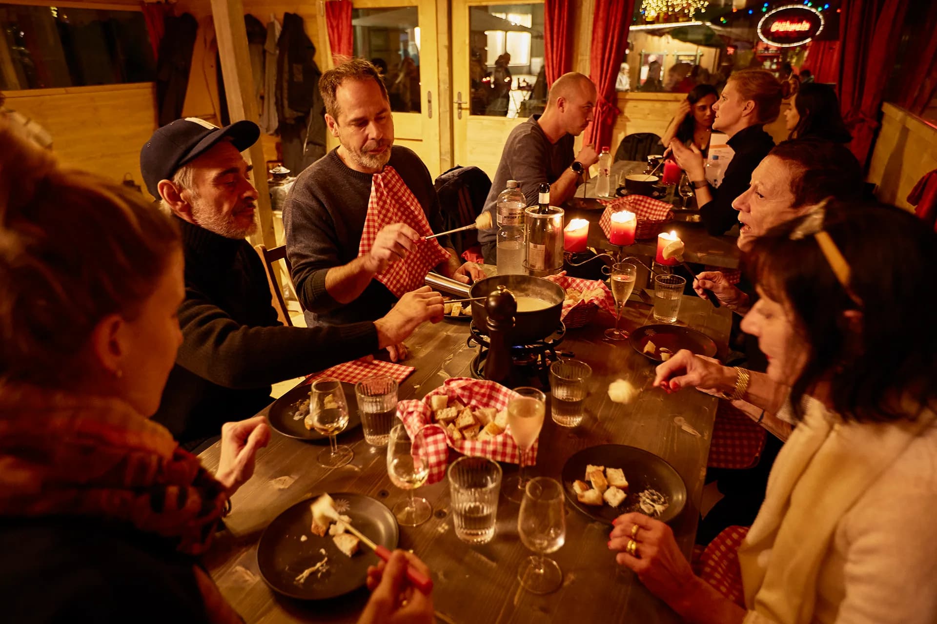 Gäste essen Käsefondue an einem langen Holztisch im Fondue-Chalet, mit Kerzen, Weingläsern und Brotkörben