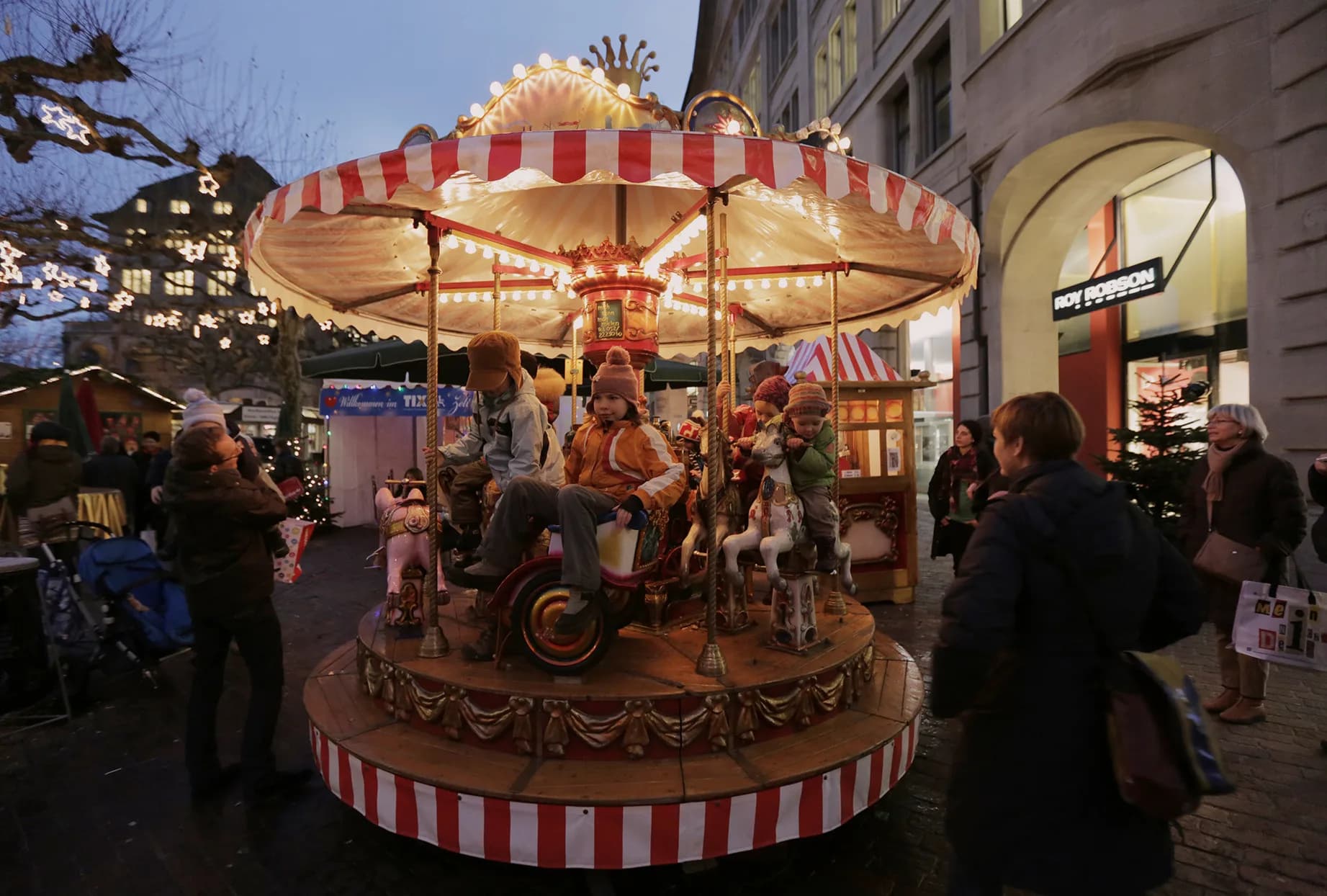 Kinder fahren auf dem rot-weiss gestreiften, beleuchteten Chinder-Karussell am Wiehnachtsmärt in der Abenddämmerung